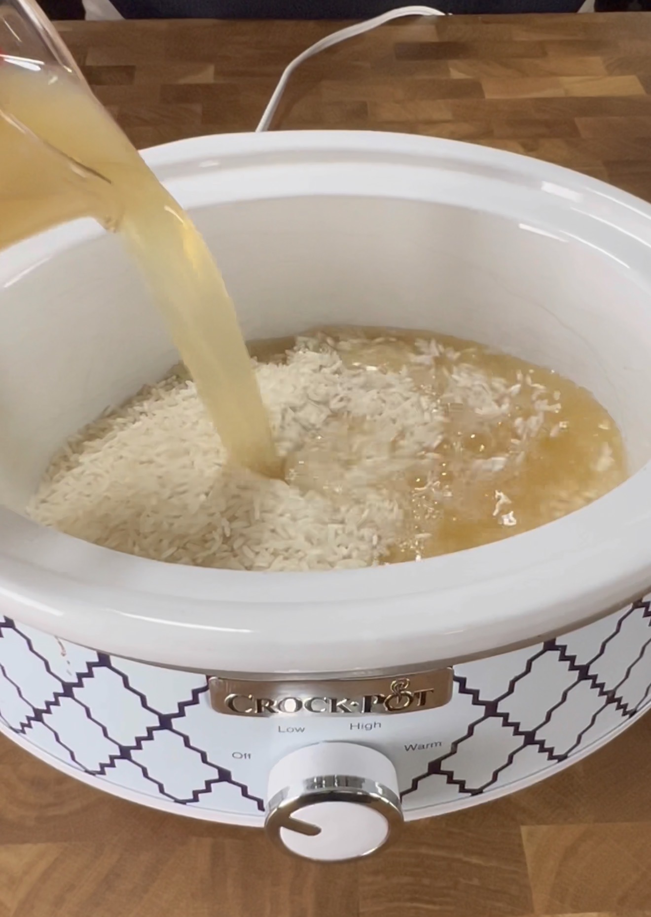 A white Crock-Pot slow cooker filled with uncooked rice, as a stream of broth or liquid is being poured in, preparing the ingredients for cooking.