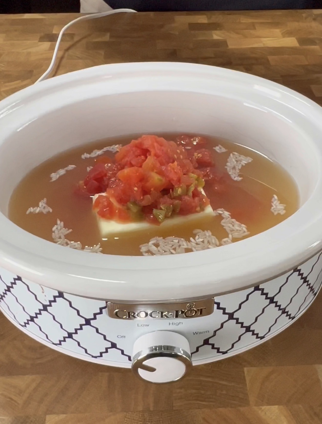 A white Crock-Pot filled with broth, uncooked rice, a block of butter, and a pile of diced tomatoes and green chilies, sitting on a wooden surface.
