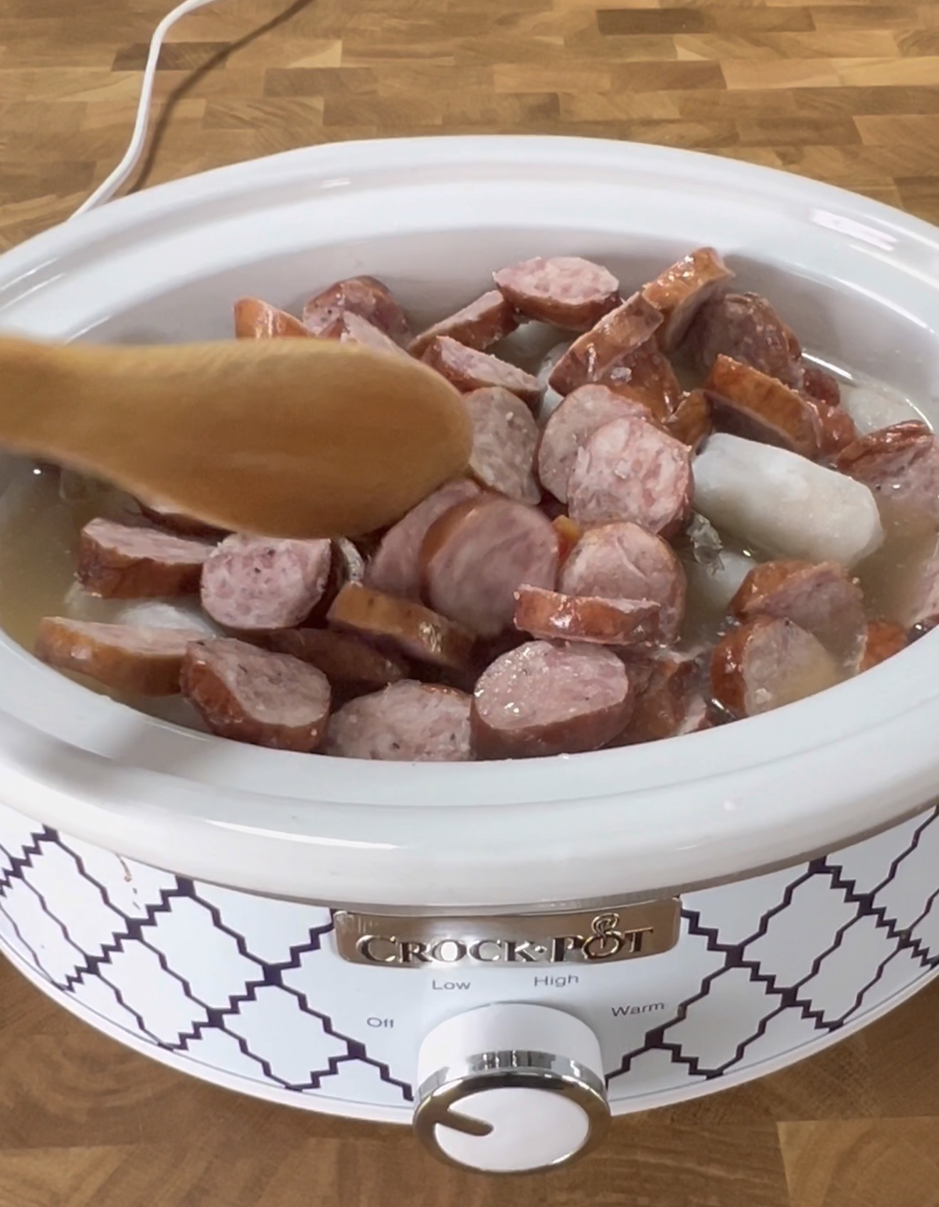 A hand stirs sliced sausage pieces in a white Crock-Pot slow cooker with a wooden spoon. The cooker is set on a wooden countertop.