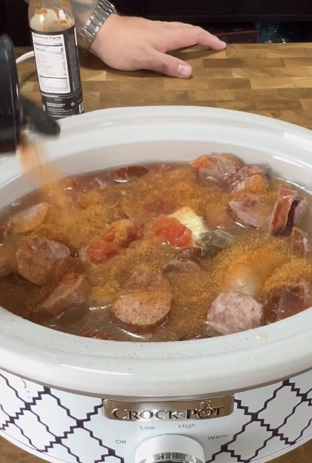 A slow cooker labeled Crock-Pot filled with sausage, tomatoes, and spices, as seasoning is sprinkled in. A persons hand and a bottle of hot sauce are visible in the background on a wooden countertop.