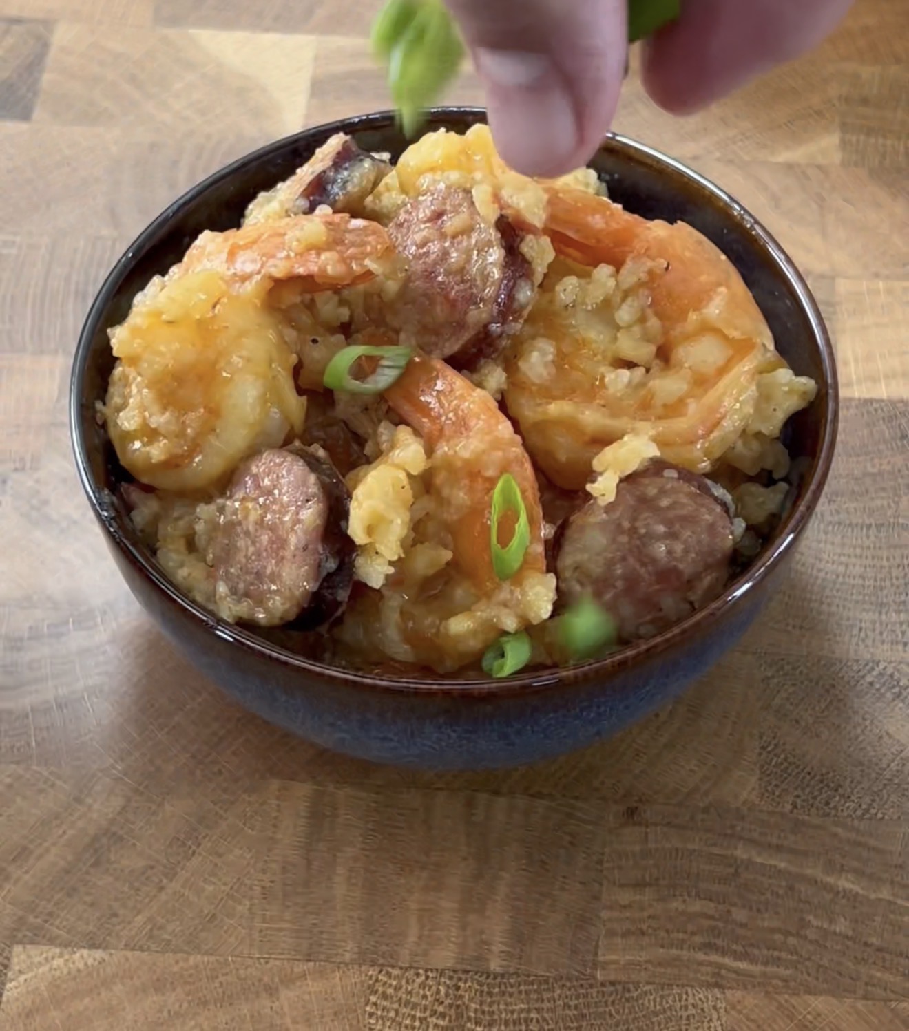 A hand sprinkles chopped green onions over a bowl of cooked shrimp and sausage on a wooden surface.