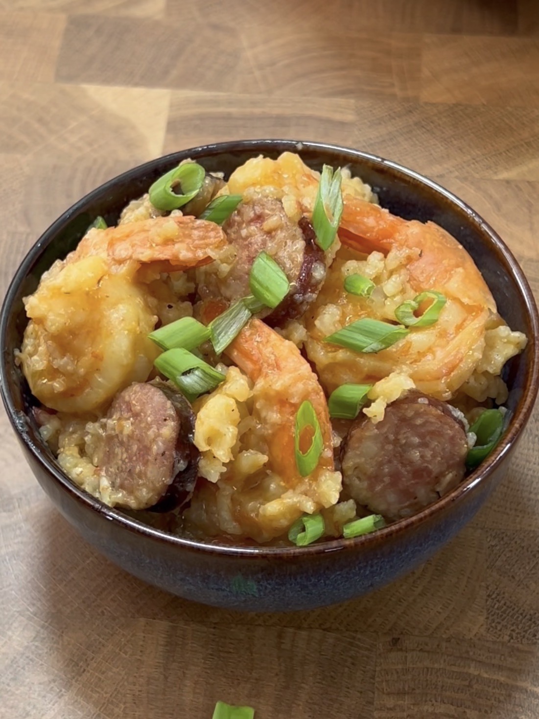 A bowl of jambalaya filled with shrimp, sausage slices, rice, and topped with chopped green onions, sitting on a wooden surface.