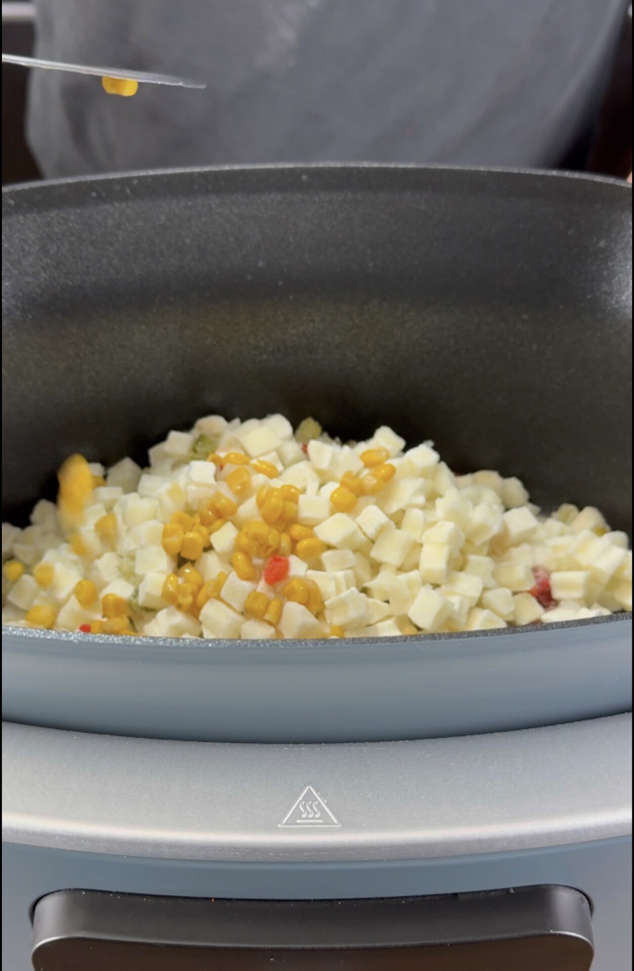 Diced vegetables, including corn and white cubes, are being cooked in a black pan on a gray stovetop. A hand above the pan holds a utensil with a small amount of orange food.