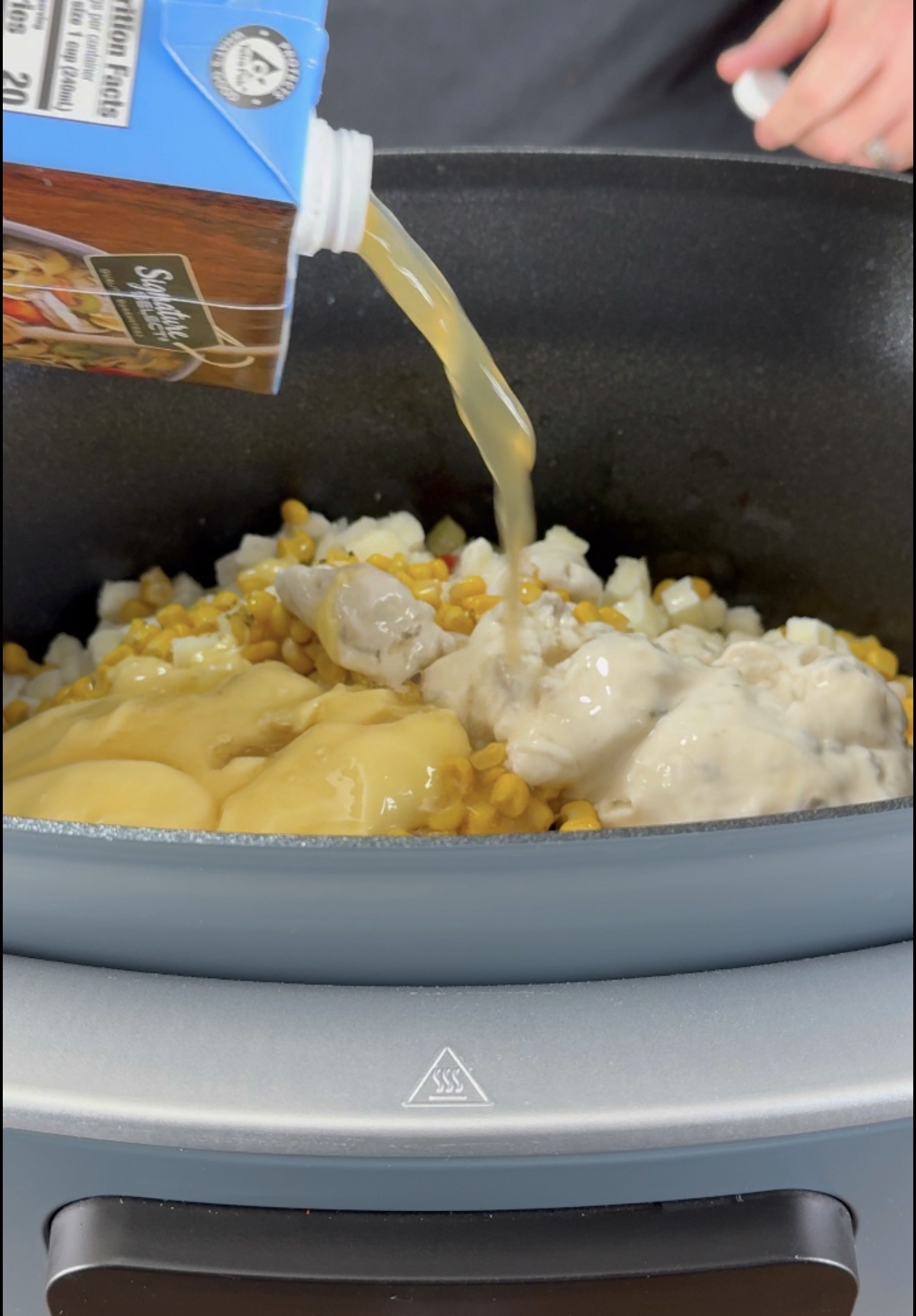 A person pours chicken broth from a carton into a slow cooker filled with corn, chopped onions, and cream soups.