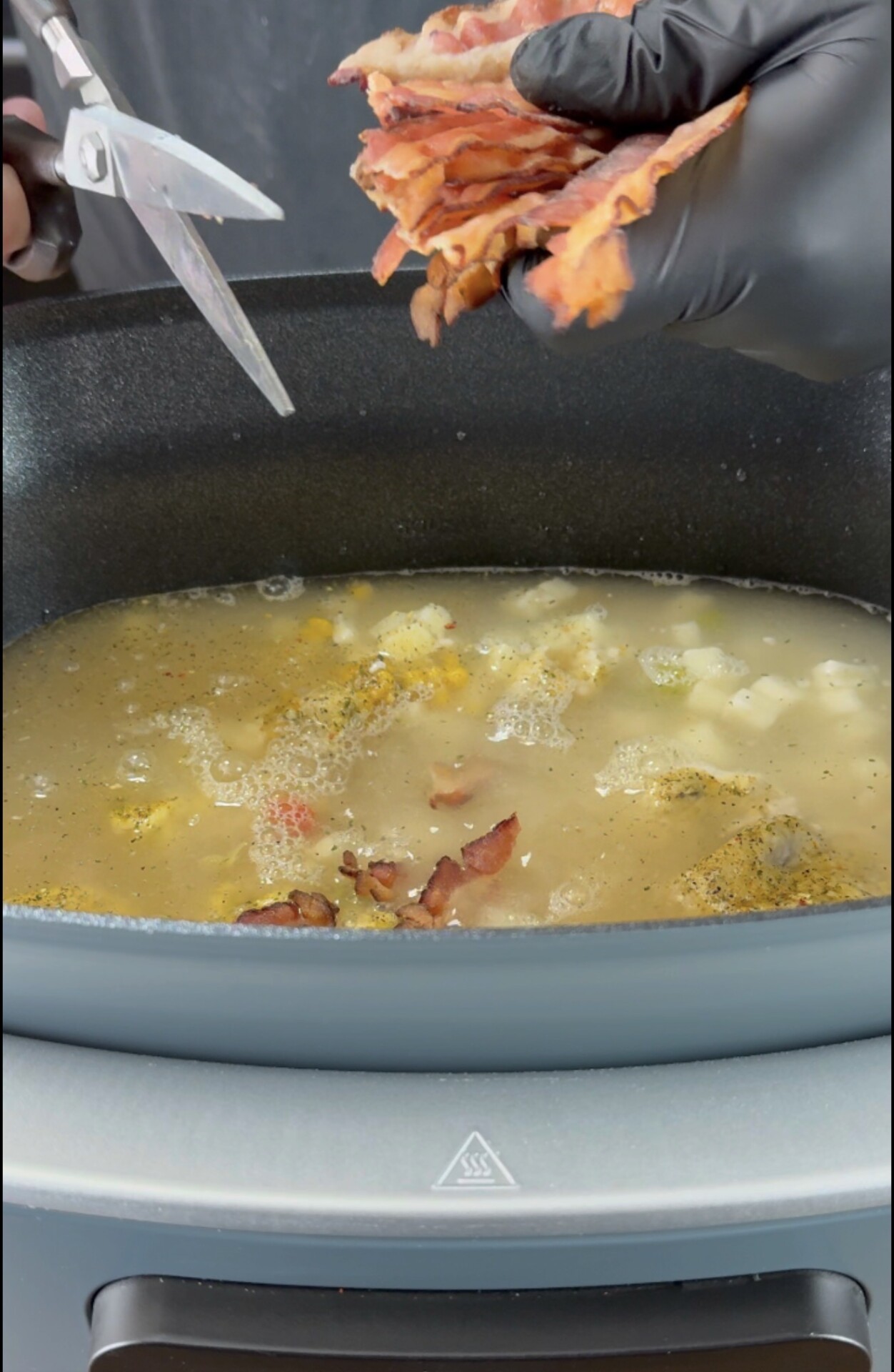 A person wearing black gloves uses scissors to cut cooked bacon into a pot of soup or stew with visible chunks of vegetables and broth.