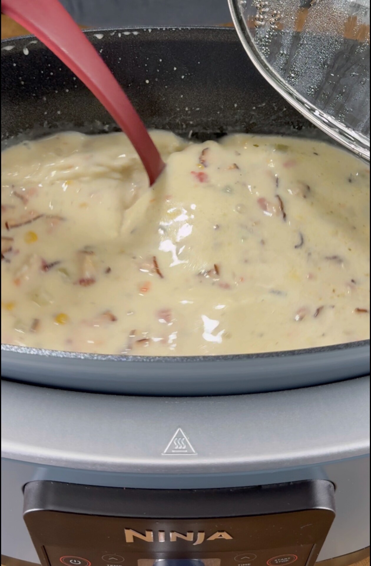 A close-up of a creamy soup with visible bits of vegetables and meat being stirred in a Ninja-brand cooking appliance using a red spatula.