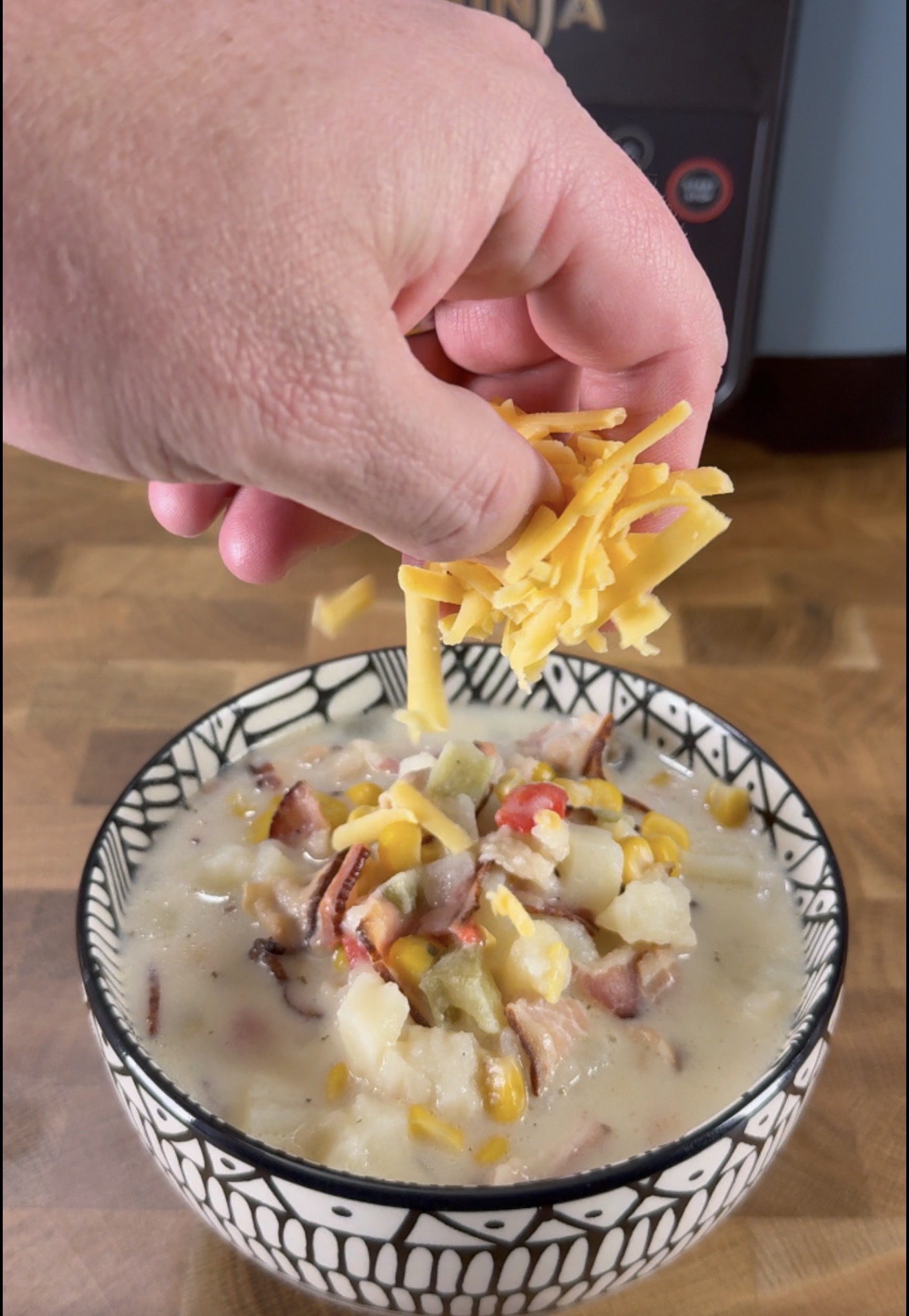 A hand sprinkles shredded cheddar cheese onto a bowl of creamy soup with corn, potatoes, and bacon on a wooden surface.