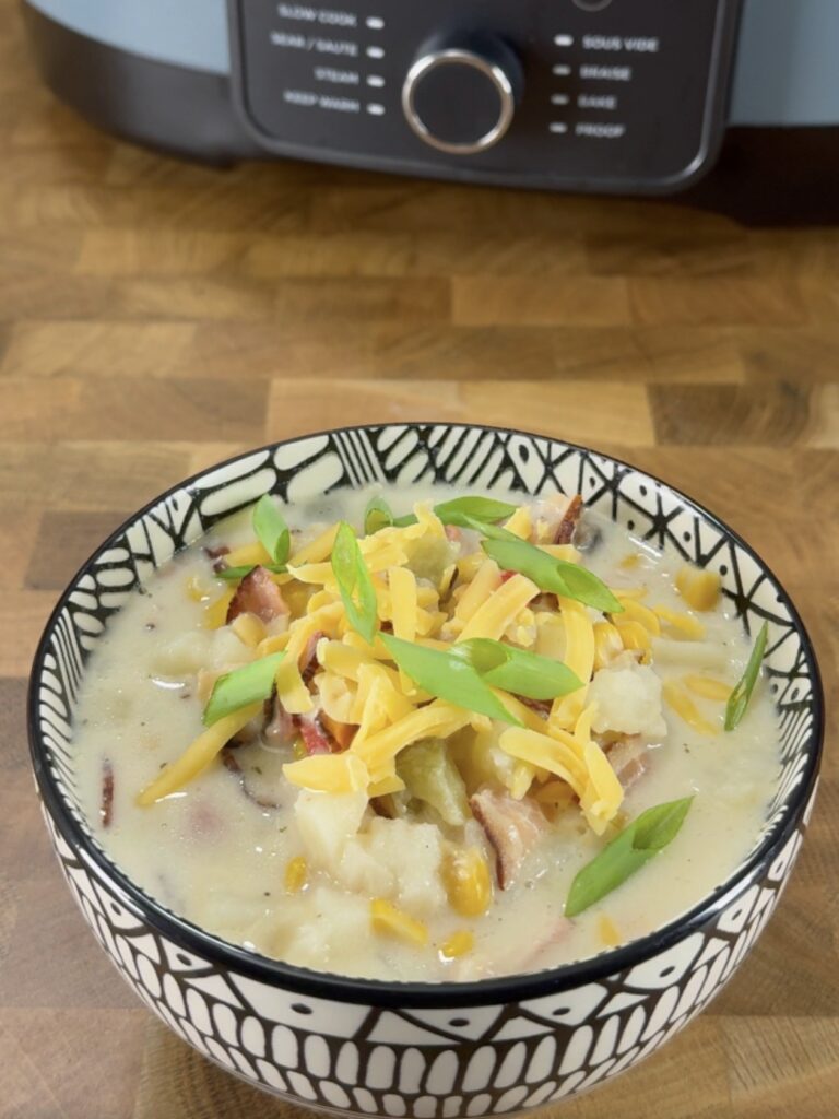 A patterned bowl filled with creamy soup, topped with shredded cheddar cheese and sliced green onions, sits on a wooden surface with a kitchen appliance in the background.