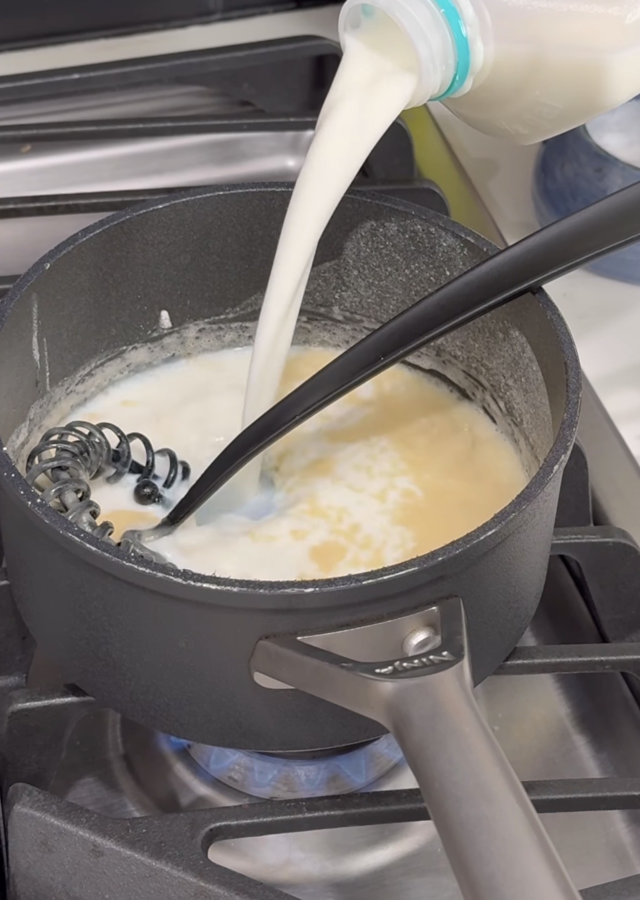 A saucepan on a stove contains a light-colored mixture being whisked, while milk is being poured in from a bottle. A black whisk and spoon rest in the pot. The burner beneath the pot is lit.