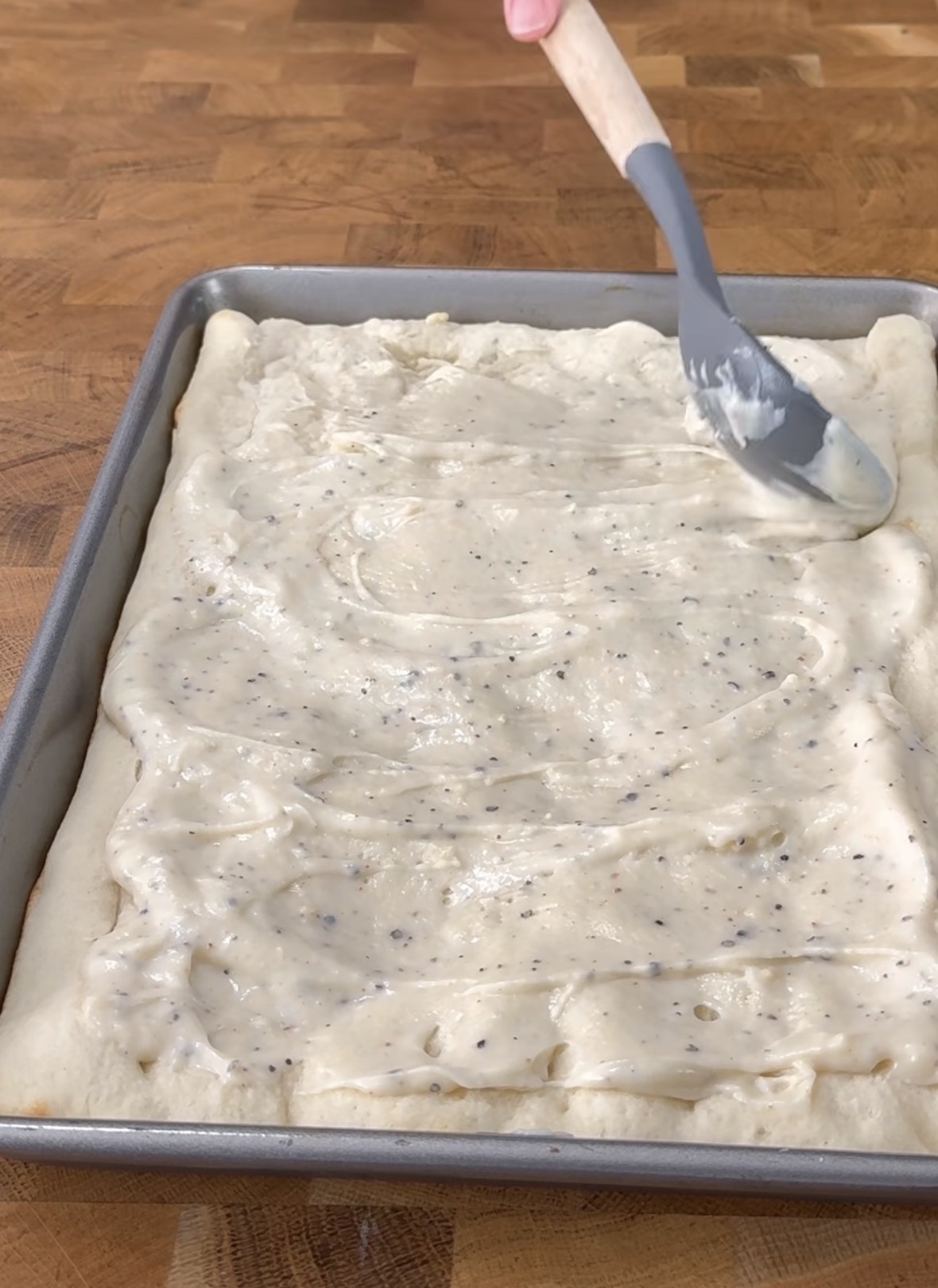 A hand uses a spatula to spread a creamy, speckled sauce over unbaked rectangular dough in a metal baking pan on a wooden surface.