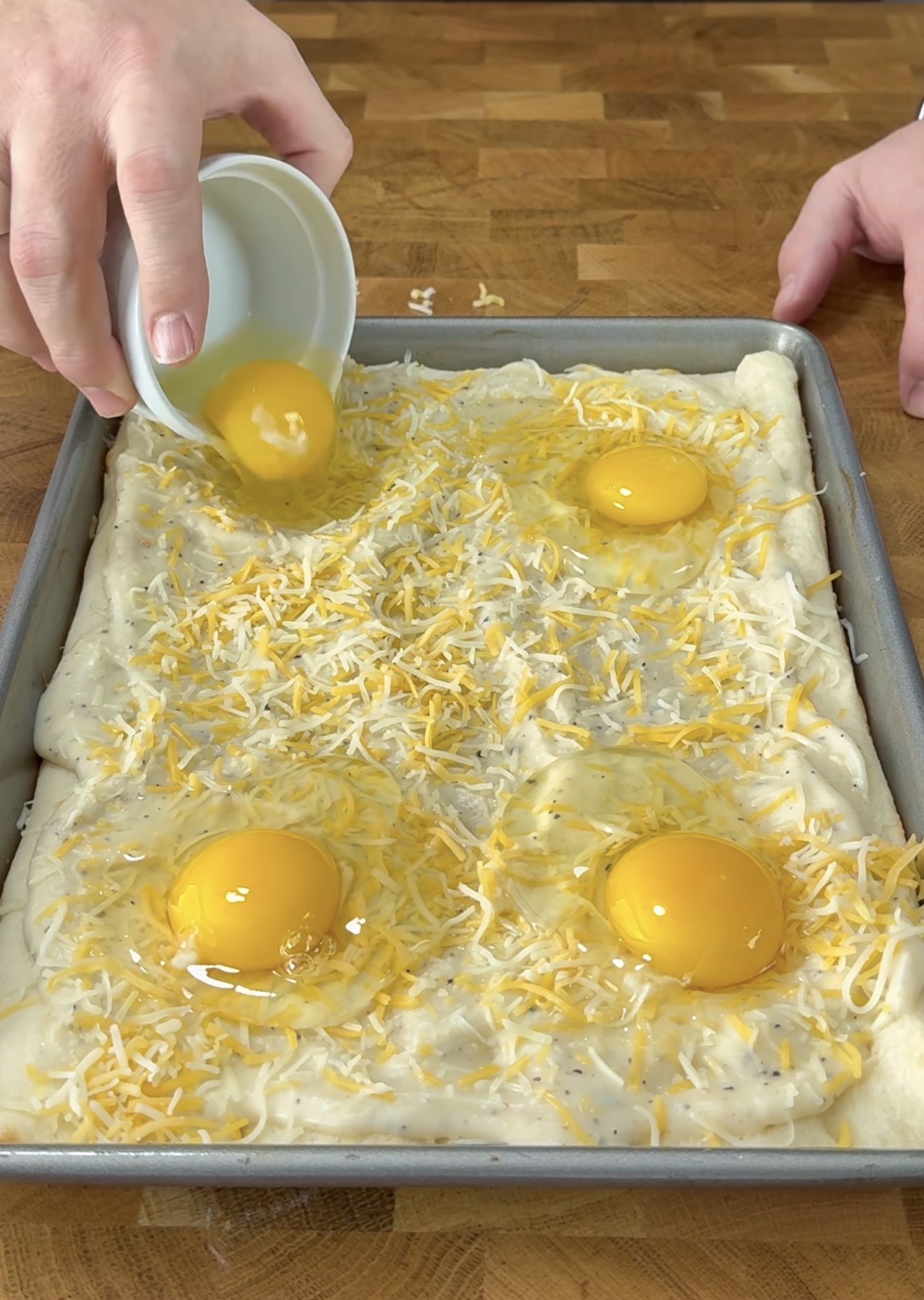 A person pours a cracked raw egg onto a rectangular sheet of dough in a baking pan, which is topped with shredded cheese and three other raw eggs, on a wooden surface.