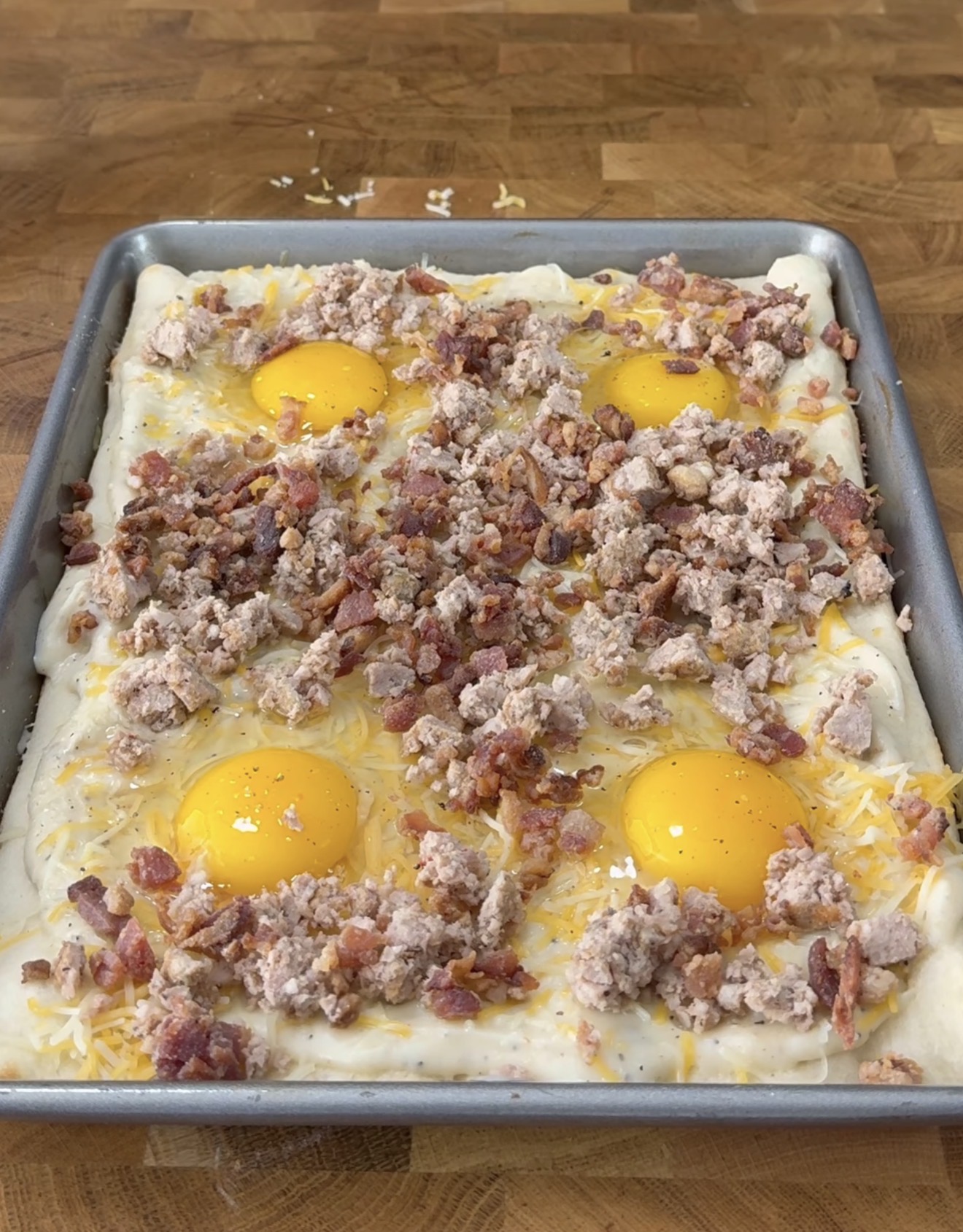 A rectangular baking pan on a wooden surface holds unbaked dough topped with four raw eggs, cooked crumbled sausage, bacon bits, and shredded cheese, arranged evenly across the surface.