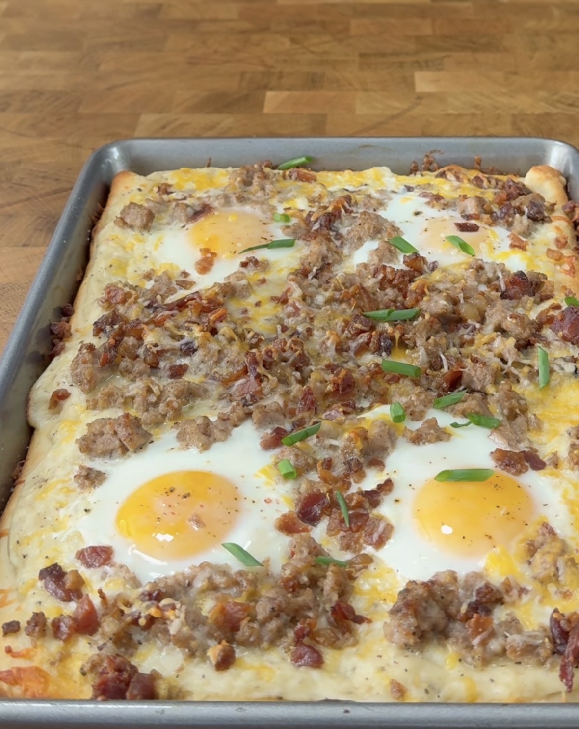 A rectangular breakfast pizza on a sheet pan, topped with melted cheese, sunny-side-up eggs, crumbled sausage, bacon pieces, and green onion slices, sits on a wooden countertop.