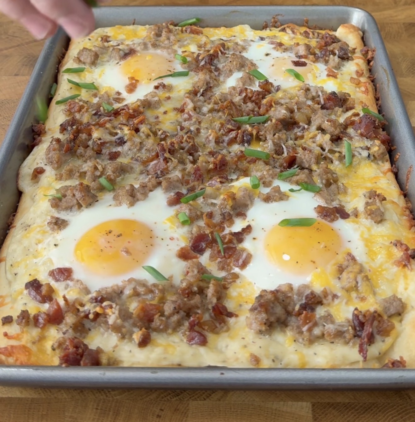 Rectangular breakfast pizza topped with sunny-side-up eggs, crumbled sausage, bacon, cheese, and chopped green onions on a metal baking sheet. A hand is visible in the top left corner.