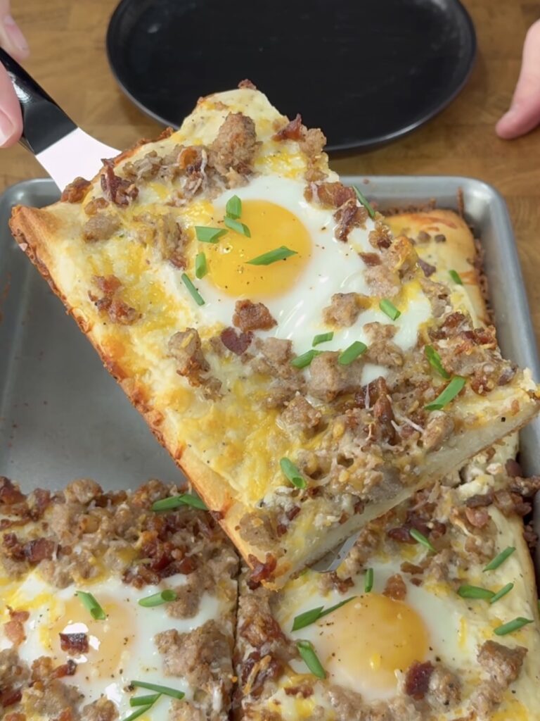A close-up of a rectangular slice of breakfast pizza topped with a sunny-side-up egg, sausage, bacon, melted cheese, and chopped chives, being lifted from a baking pan.