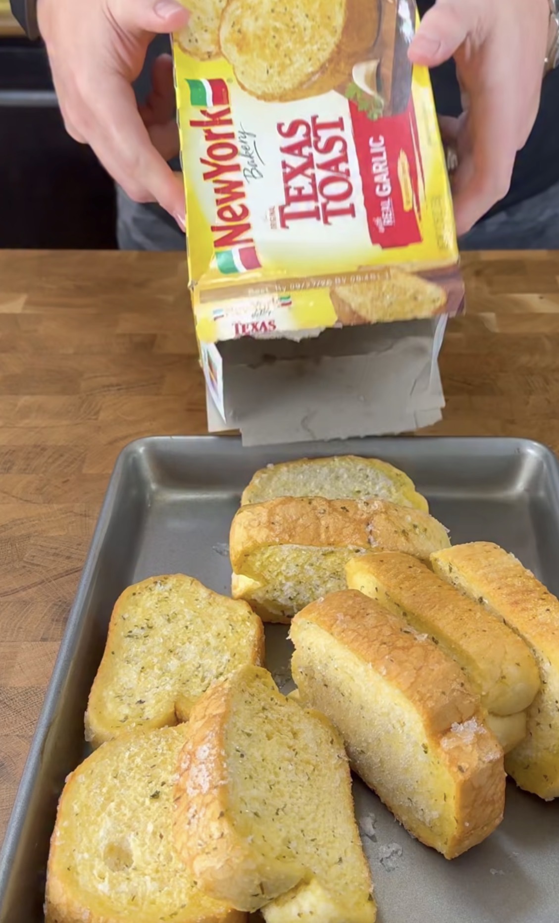 A person places slices of New York Bakery Texas Toast with garlic on a baking tray, with the open yellow box in their hands above the tray.