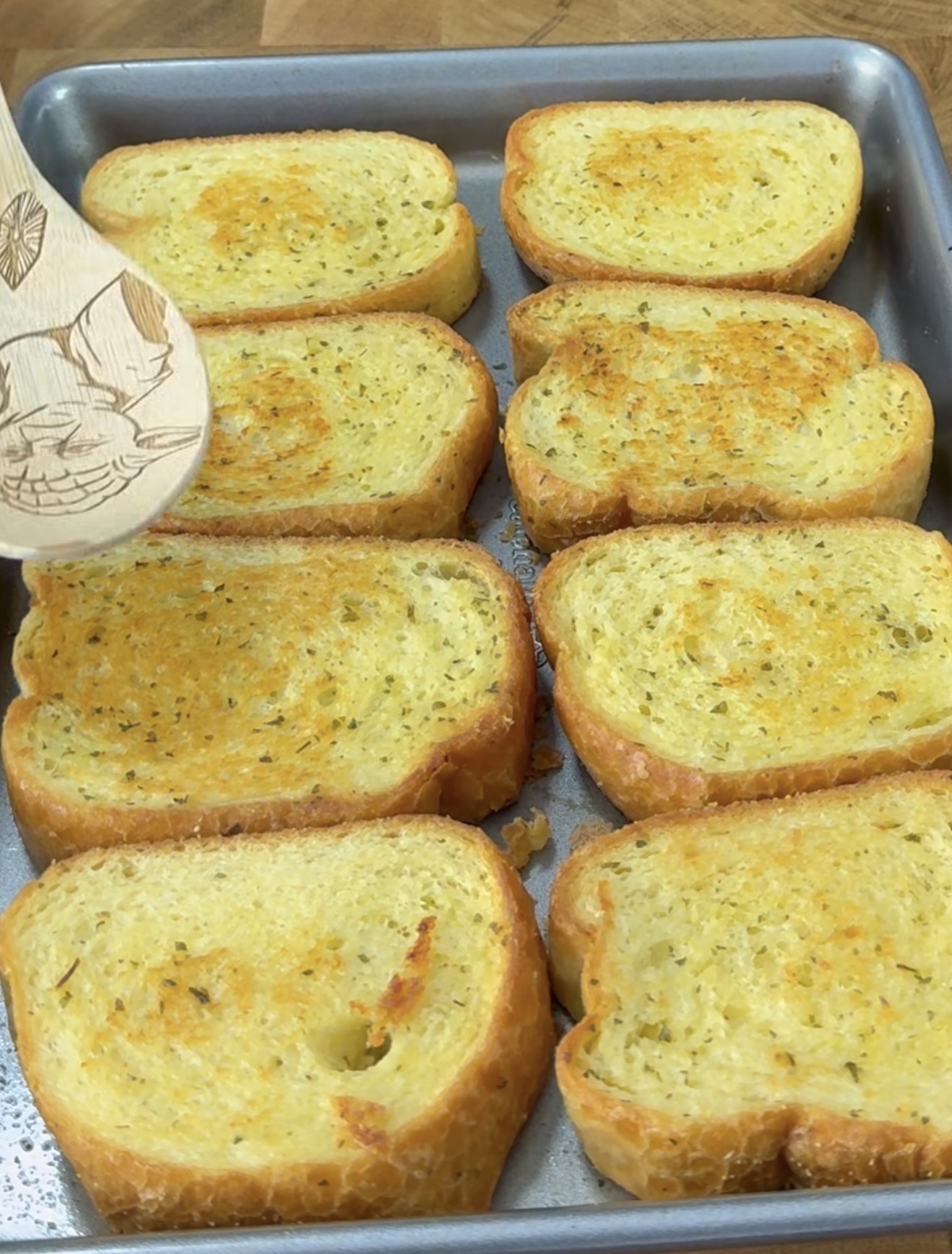 Eight slices of golden, toasted garlic bread are arranged on a baking tray. A wooden spatula is lifting one slice, revealing the crispy texture and seasoning on the bread.