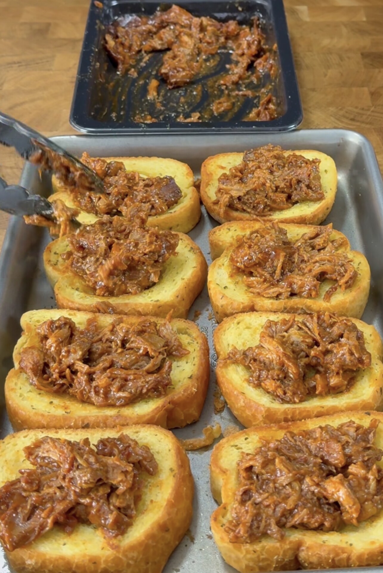Slices of toasted bread on a baking tray are topped with shredded, saucy barbecue meat using tongs. More of the meat mixture sits in a tray in the background on a wooden surface.