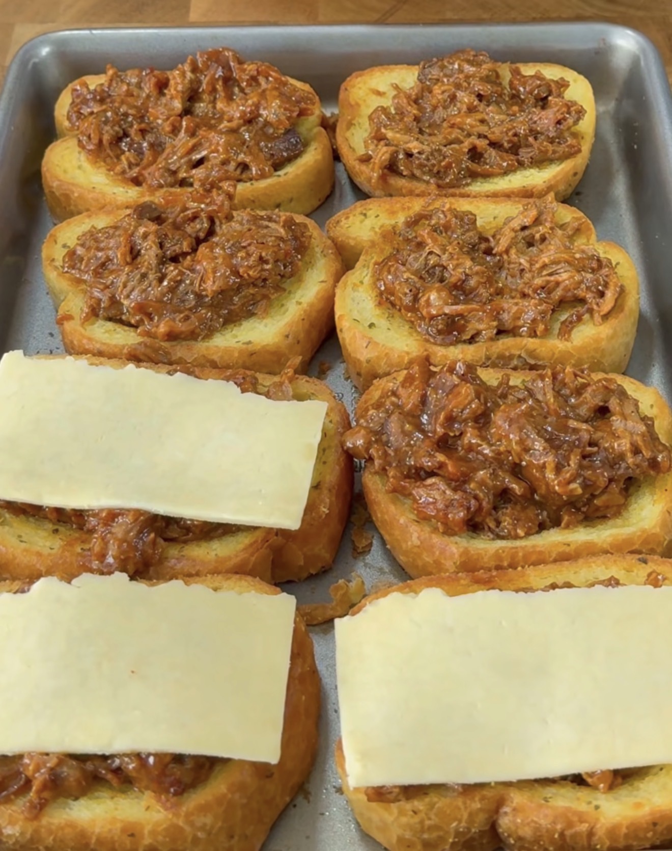 Eight slices of bread on a baking tray, topped with a shredded meat mixture; three slices also have a piece of cheese placed on top of the meat.