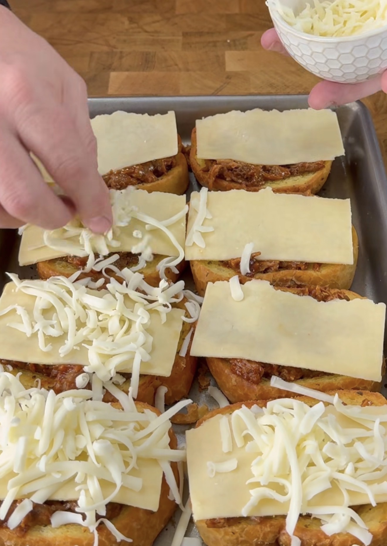 A hand sprinkles shredded cheese onto open-faced sandwiches topped with sliced cheese and meat sauce, all arranged on a baking tray.