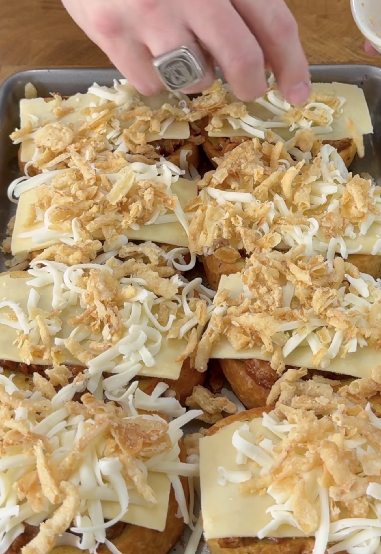 A hand sprinkles shredded cheese and crispy fried onions over a tray of sliced bread topped with cheese, preparing a baked dish.