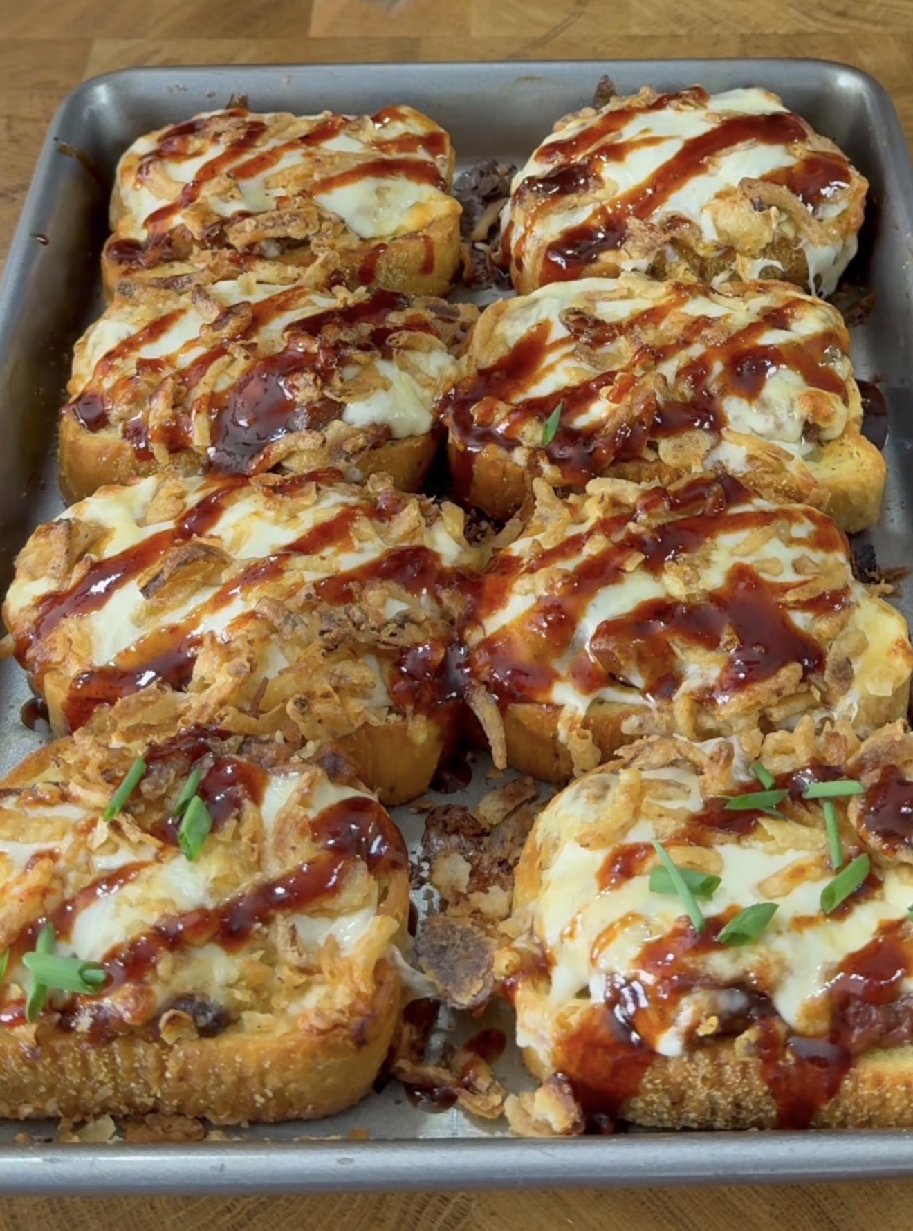 A baking tray filled with nine pieces of toasted bread topped with melted cheese, crispy fried onions, barbecue sauce, and garnished with chopped green herbs.