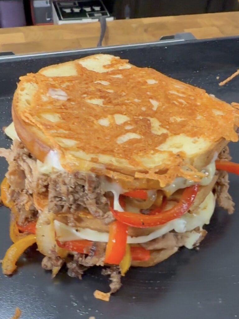 A close-up of a hearty sandwich with melted cheese, shredded beef, and sliced red and yellow bell peppers between two thick slices of toasted bread. The sandwich appears hot and freshly made.