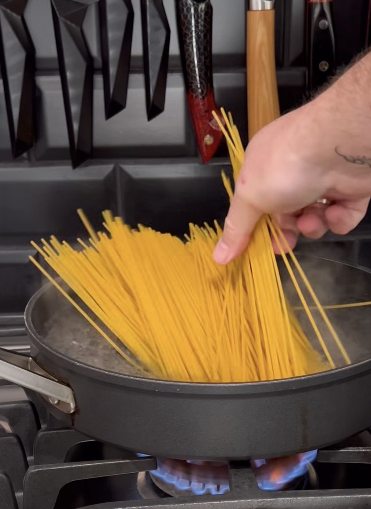 A hand adds uncooked spaghetti into a boiling pot of water on a gas stove, with several kitchen knives hanging in the background.