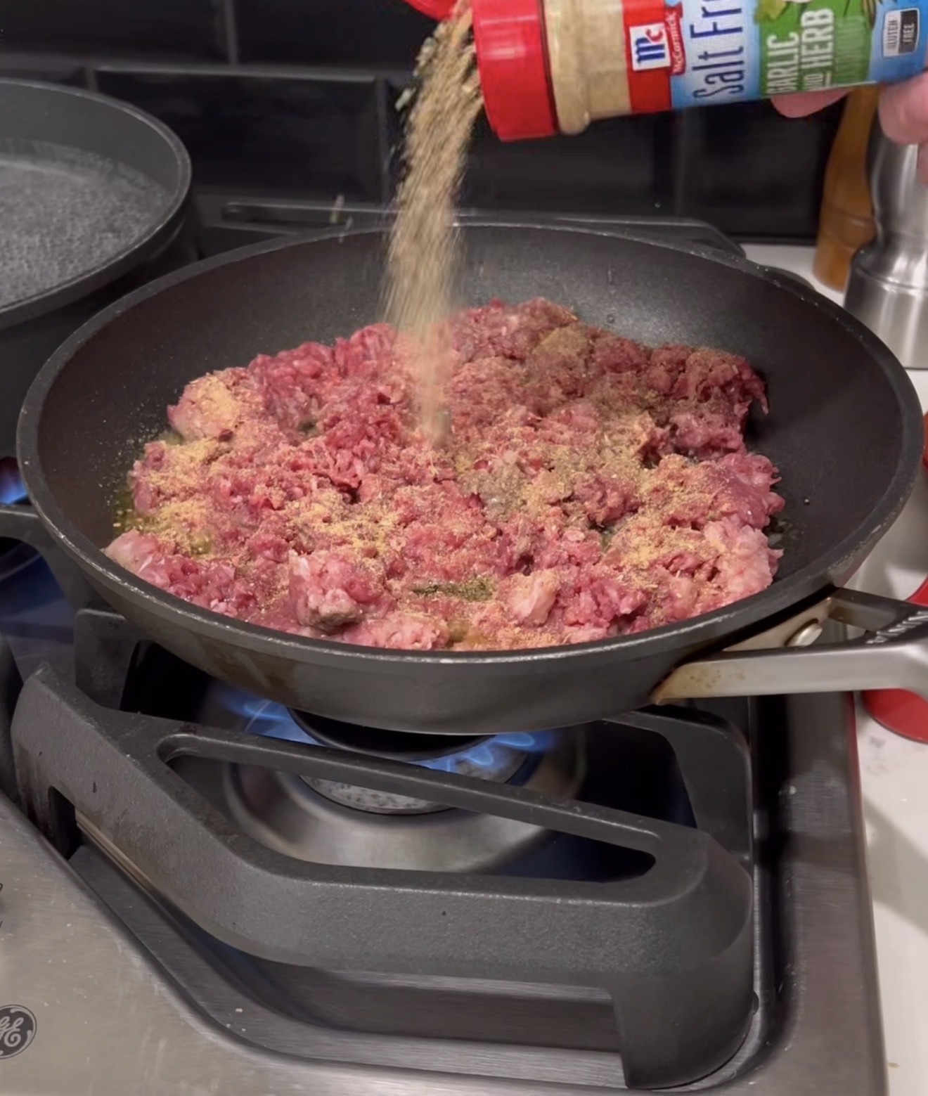 Ground beef cooking in a black skillet on a stove, with seasoning being sprinkled from a container labeled “Salt Free Garlic & Herb” over the meat. A pot of water sits on the back burner.