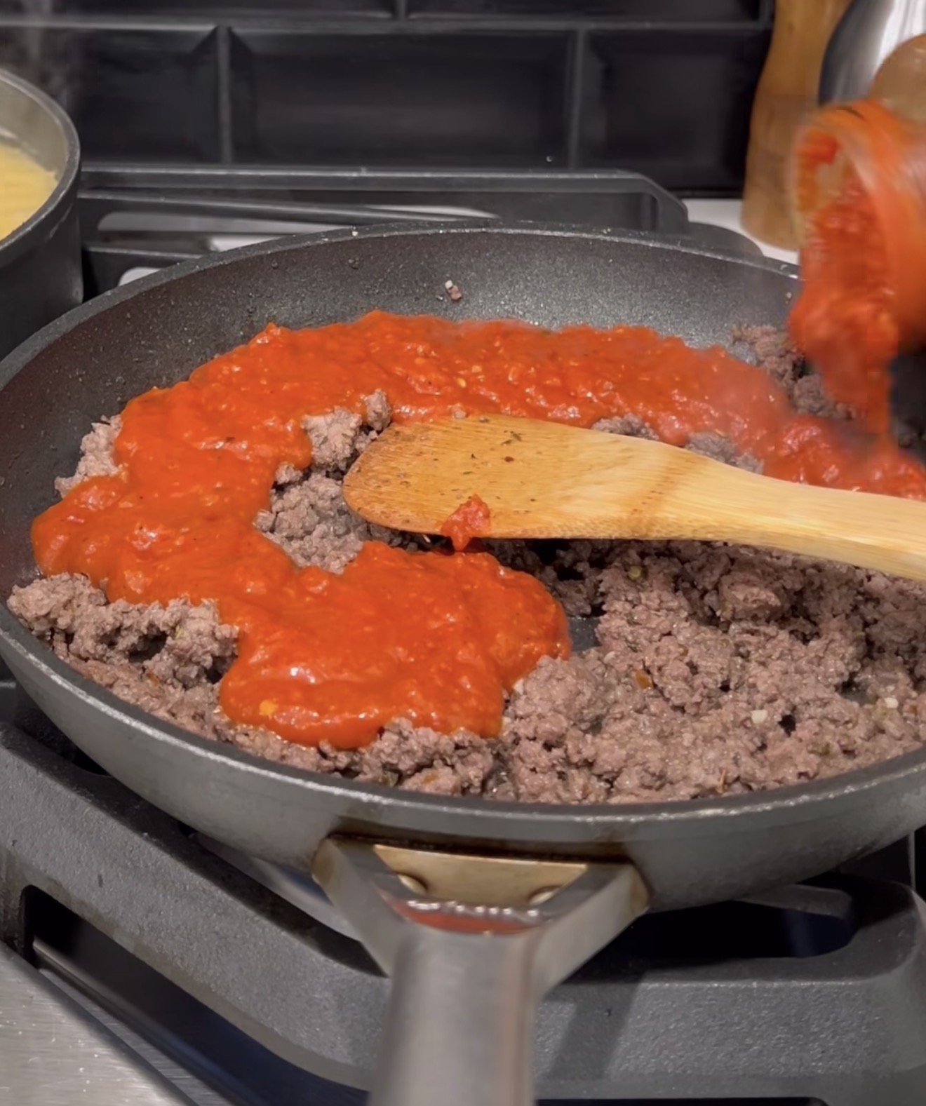 A skillet on a stovetop with cooked ground beef being mixed with tomato sauce using a wooden spatula. A pot of boiling pasta is visible in the background.