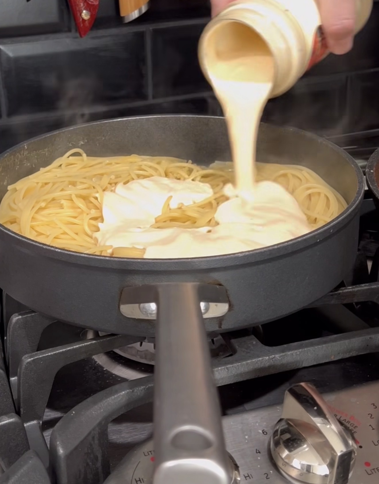 A hand pours creamy white sauce from a jar onto cooked spaghetti in a black pan on a stovetop with the burner on.