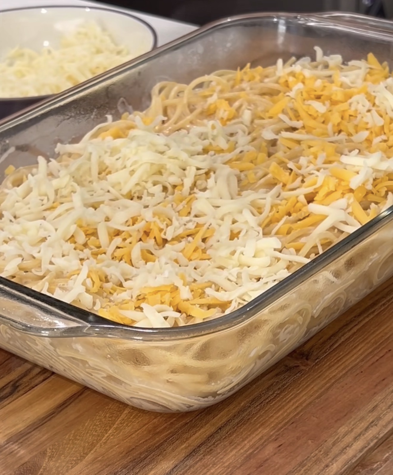A glass baking dish filled with uncooked spaghetti mixed in a creamy sauce, topped with a generous layer of shredded yellow and white cheese, sits on a wooden surface. A bowl of extra shredded cheese is nearby.