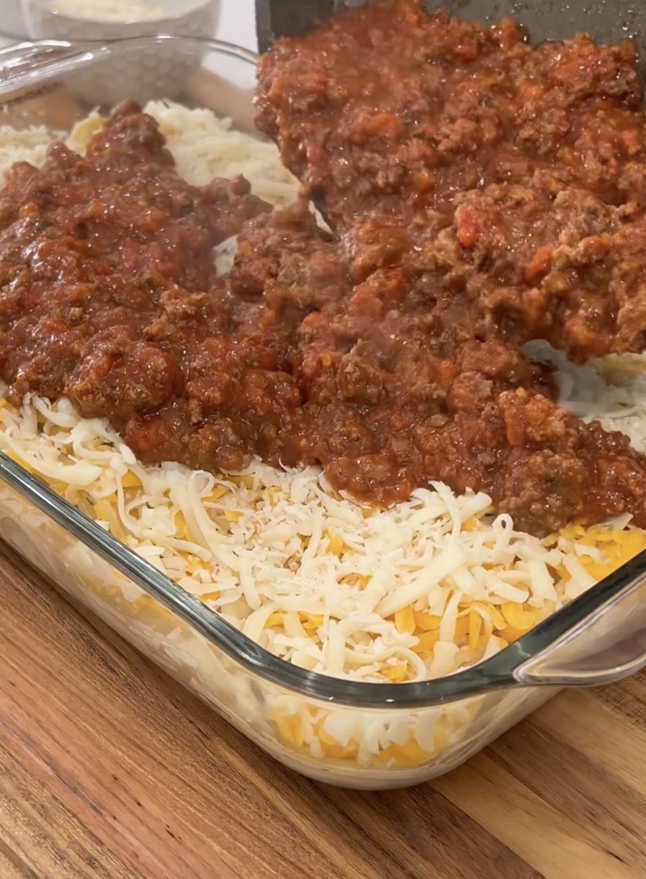 A glass baking dish is filled with shredded cheese and cooked pasta, while a layer of meat and tomato sauce is being spread on top. The dish sits on a wooden countertop.