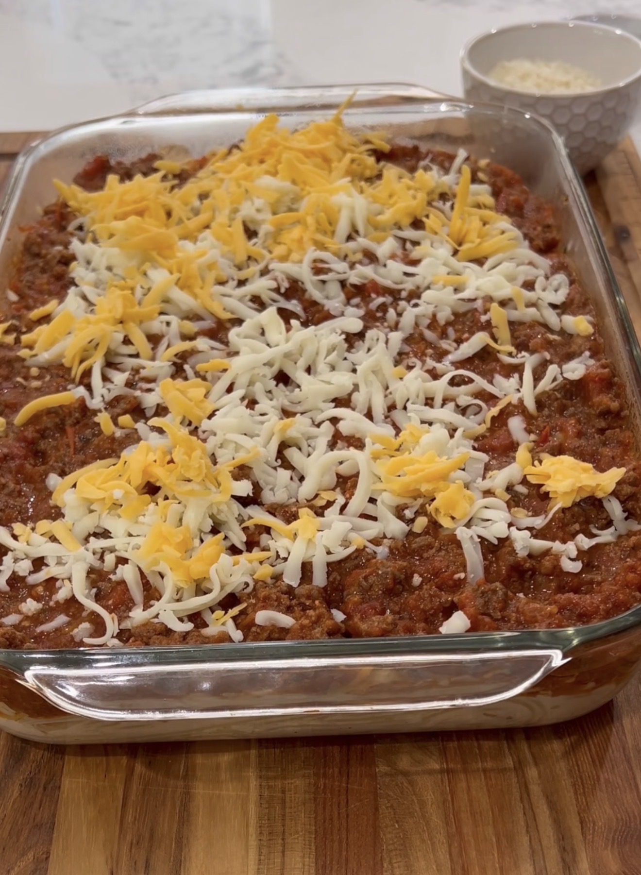 A glass baking dish filled with a layered casserole topped with shredded cheddar and mozzarella cheese, set on a wooden surface. A small bowl of extra shredded cheese is in the background.