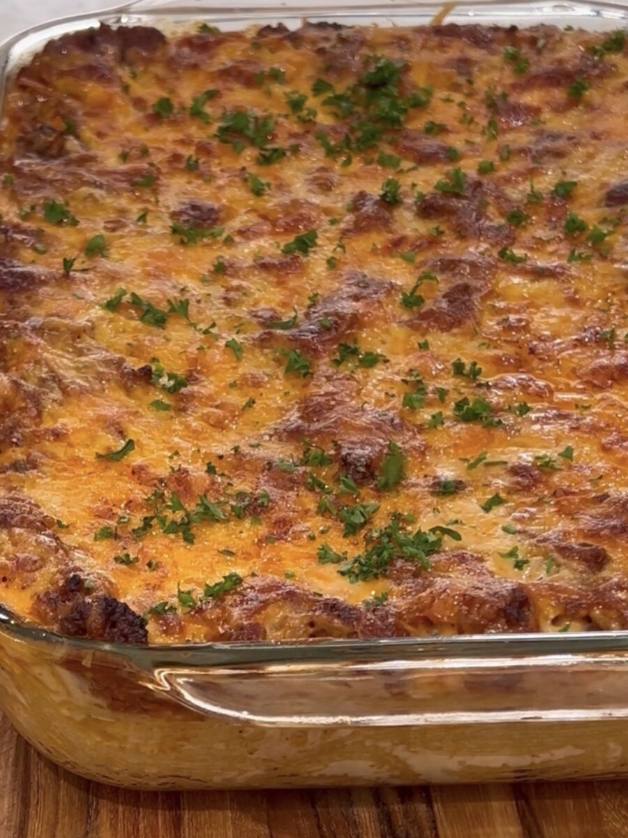 A close-up of a baked casserole in a glass dish, topped with melted, golden-brown cheese and sprinkled with chopped fresh parsley. The casserole sits on a wooden surface.