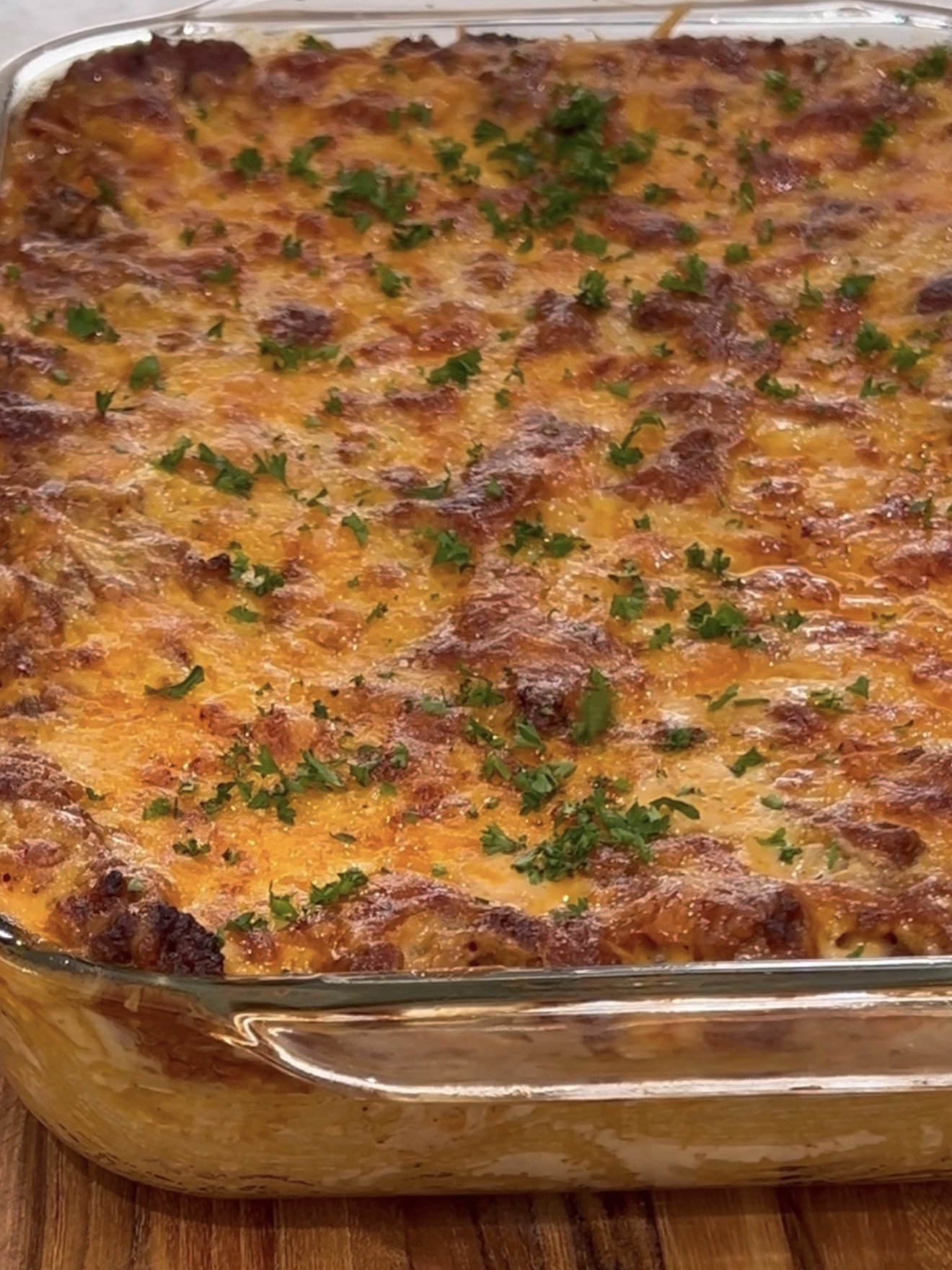 A close-up of a baked casserole in a glass dish, topped with melted, golden-brown cheese and sprinkled with chopped fresh parsley. The casserole sits on a wooden surface.
