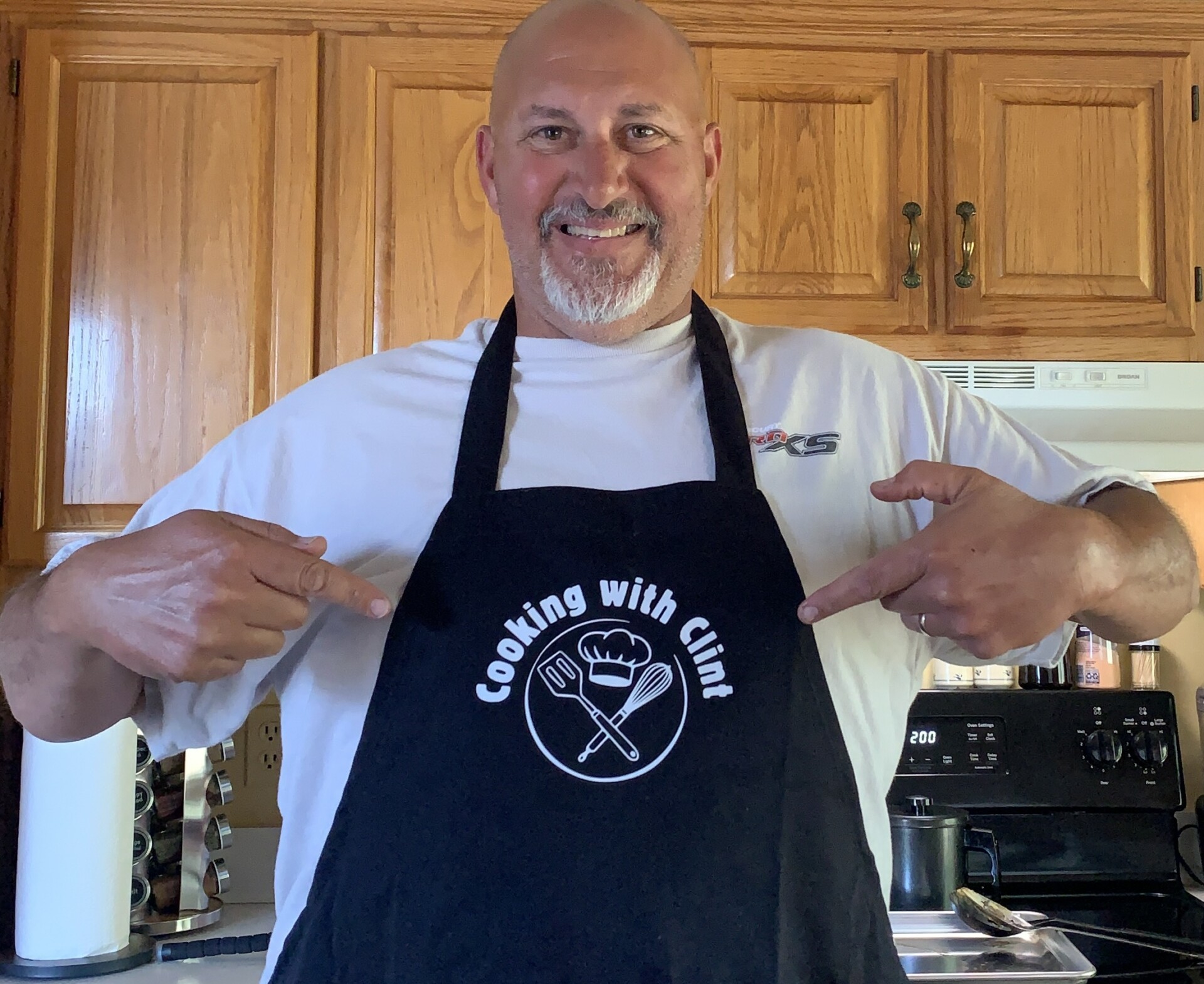 A smiling man stands in a kitchen wearing a black apron that says Cooking with Clint with cooking utensil graphics, pointing to the text on the apron.
