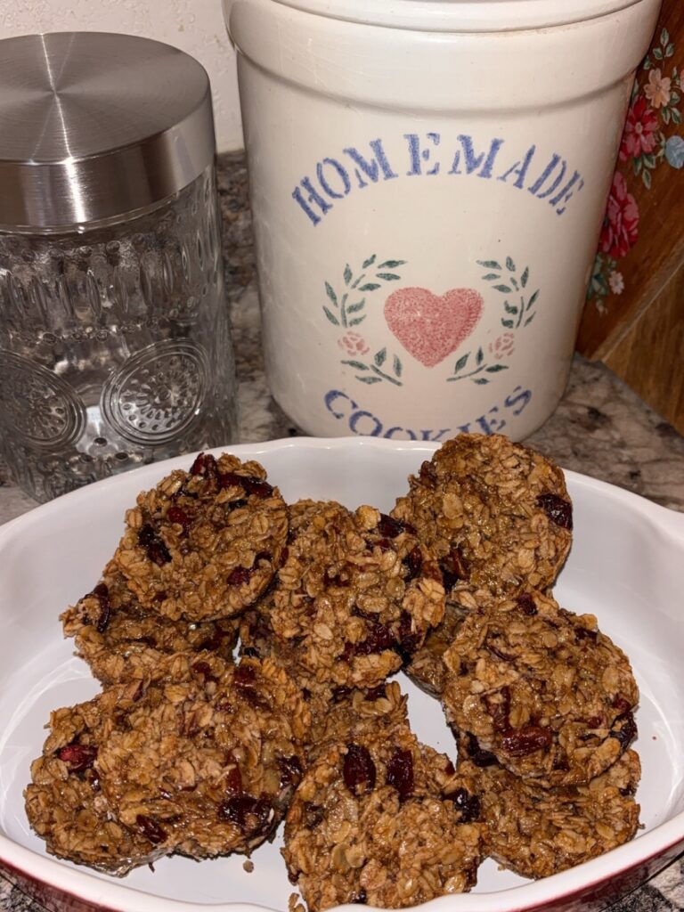 A white dish holds a batch of homemade oatmeal cookies with visible cranberries, placed on a kitchen counter beside a glass jar and a large ceramic cookie jar labeled Homemade Cookies with a pink heart and floral design.