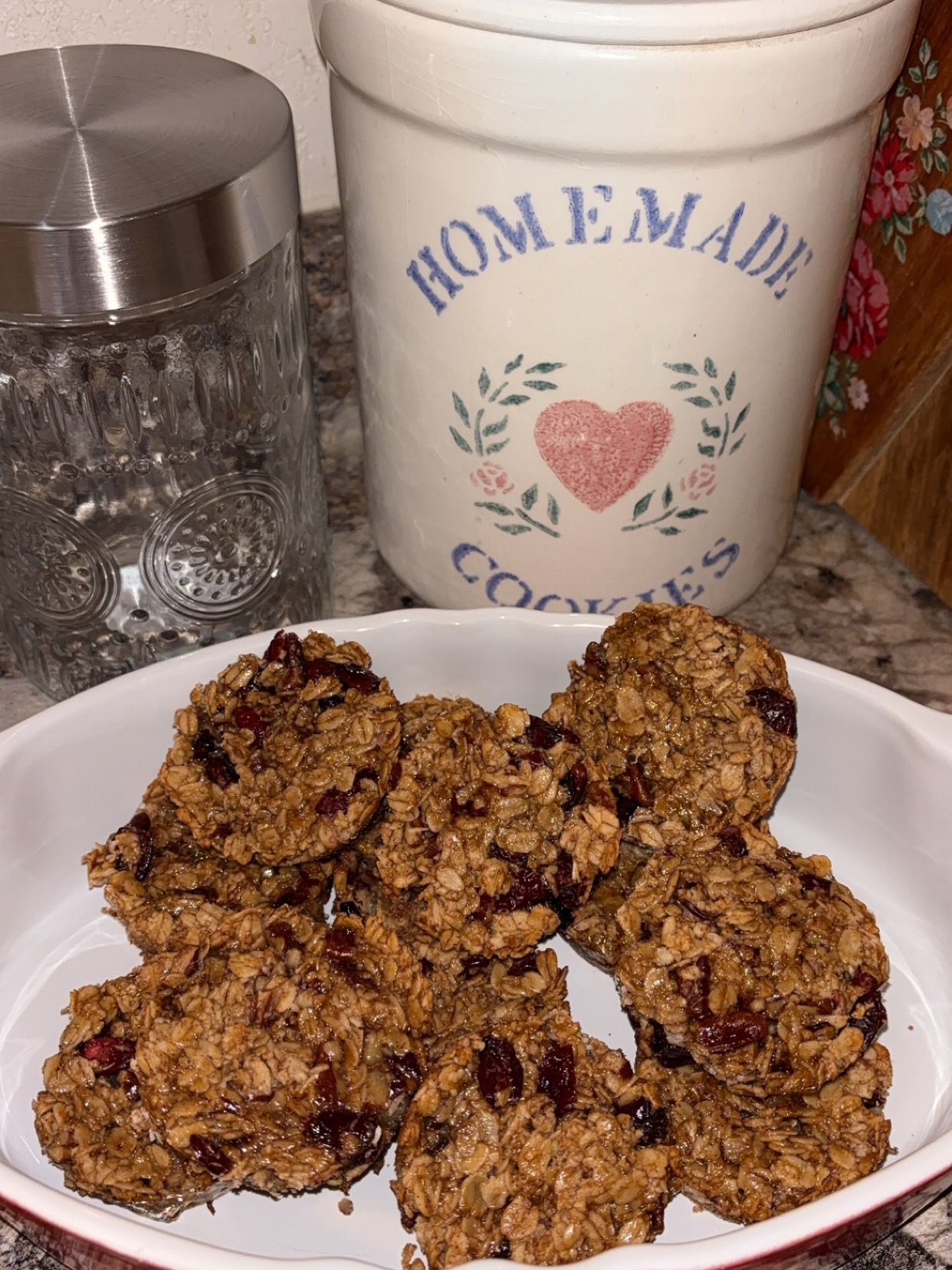 A white dish holds a batch of homemade oatmeal cookies with visible cranberries, placed on a kitchen counter beside a glass jar and a large ceramic cookie jar labeled Homemade Cookies with a pink heart and floral design.