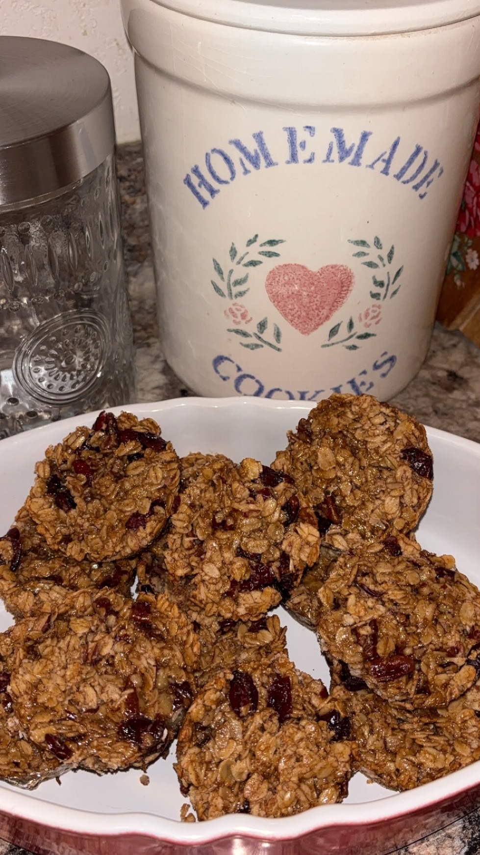 A plate of homemade oatmeal cookies with cranberries sits on a counter. Behind the plate are a glass jar and a white ceramic container labeled HOMEMADE COOKIES with a pink heart and floral design.