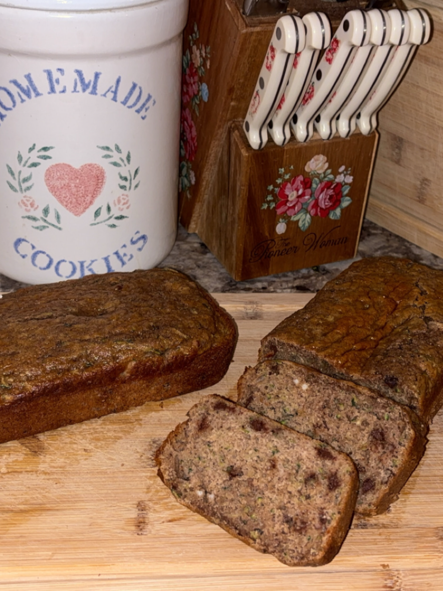 Two loaves of chocolate chip zucchini bread on a wooden cutting board, with one loaf sliced. In the background are a Homemade Cookies jar and a floral knife block on a kitchen counter.