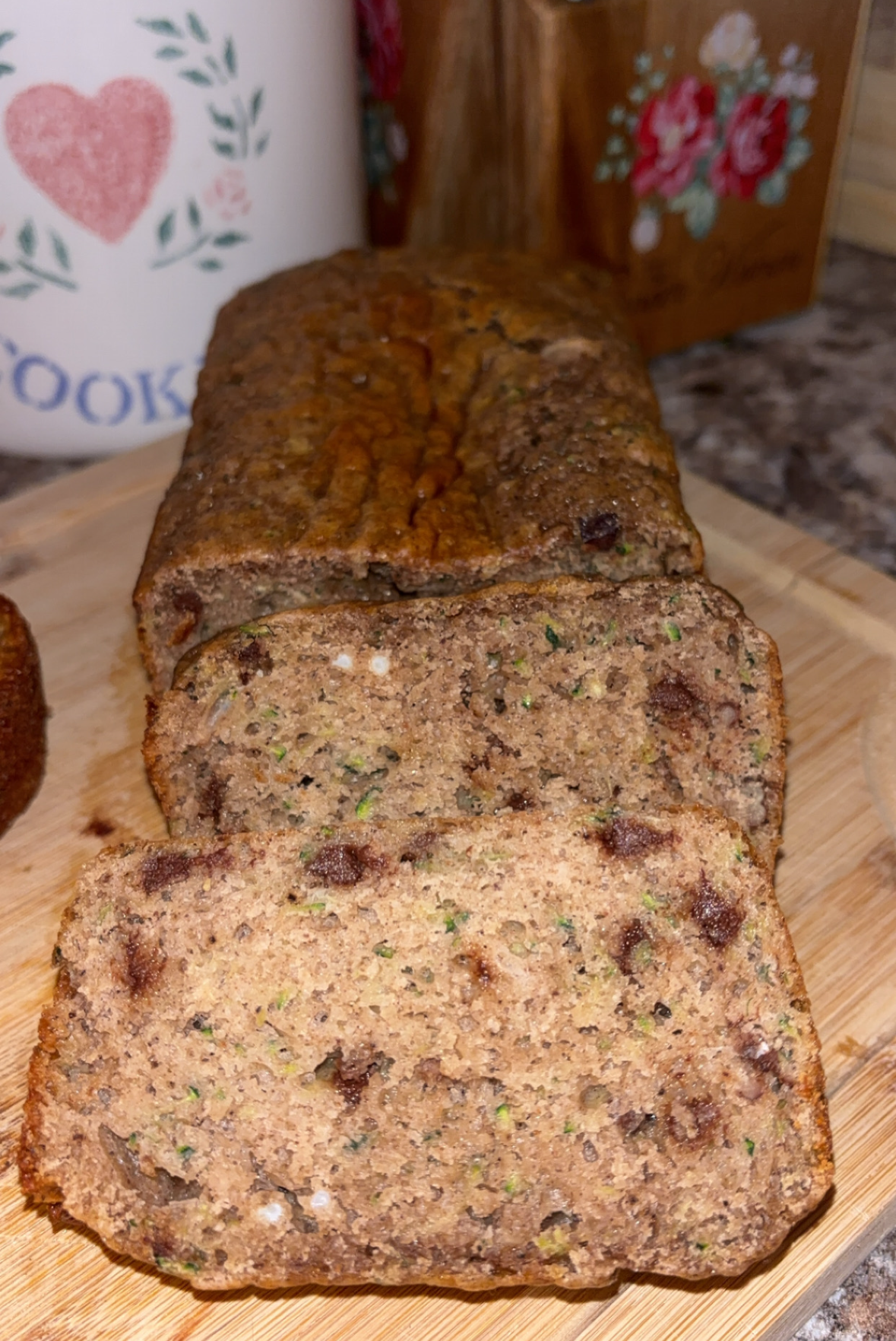 A loaf of zucchini bread with chocolate chips, partially sliced, sits on a wooden cutting board in a kitchen setting. The moist texture and visible bits of zucchini and chocolate are shown in each slice.