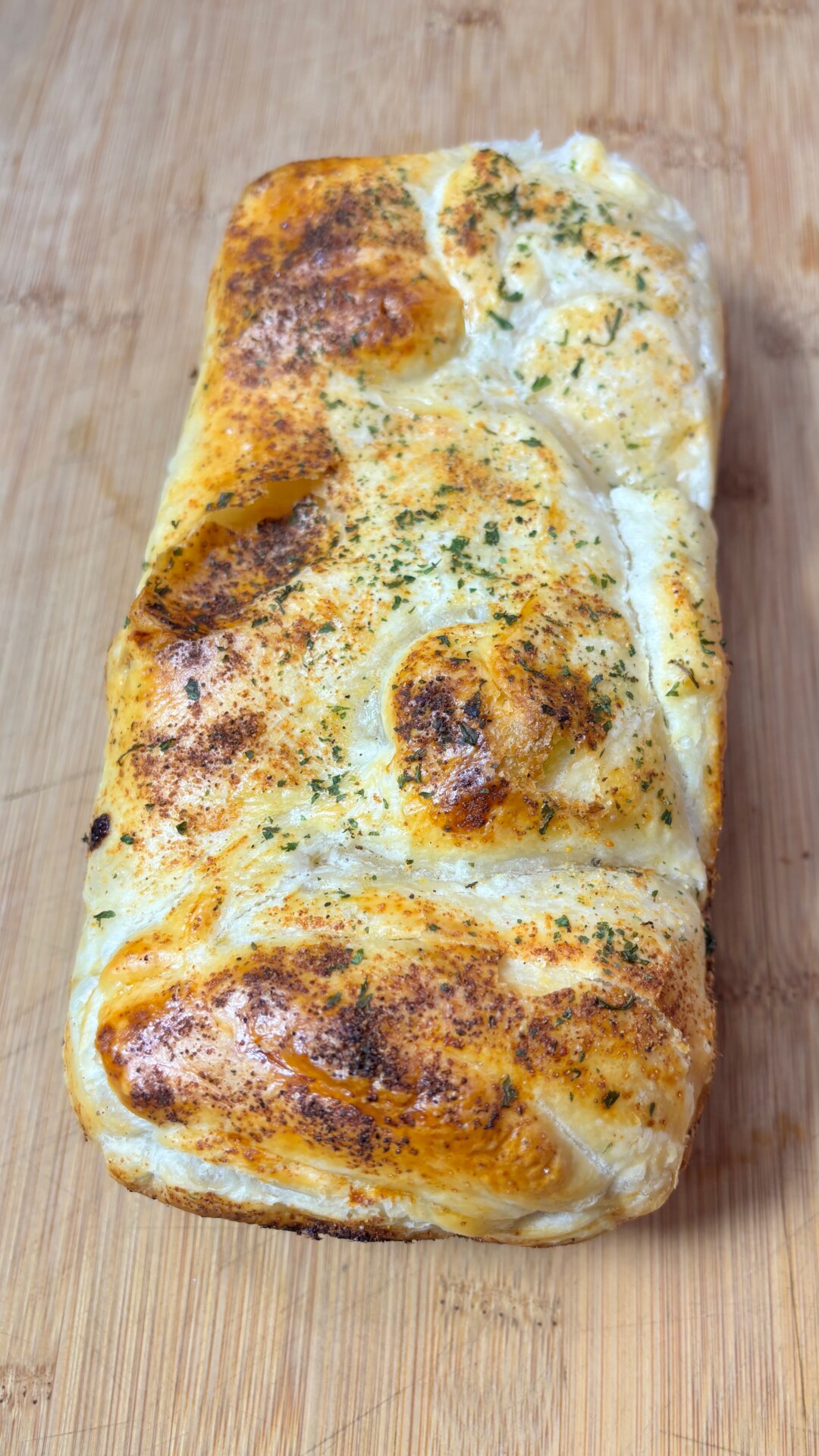 A rectangular loaf of baked bread with a golden-brown crust, sprinkled with green herbs and some darker browned spots, resting on a wooden surface.