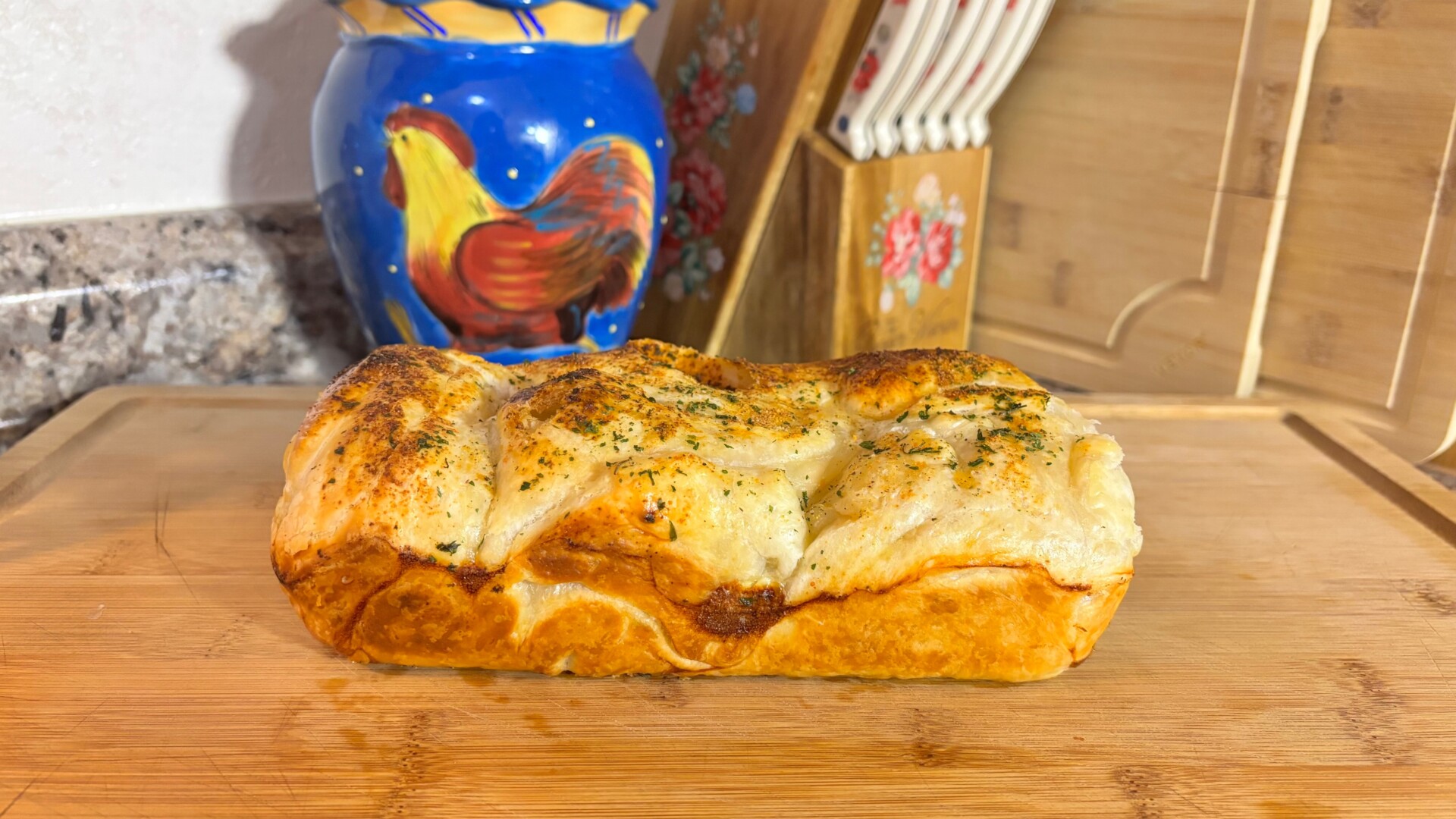 A golden-brown loaf of bread topped with herbs sits on a wooden cutting board in a kitchen, with a blue ceramic jar featuring a painted rooster and a knife block in the background.