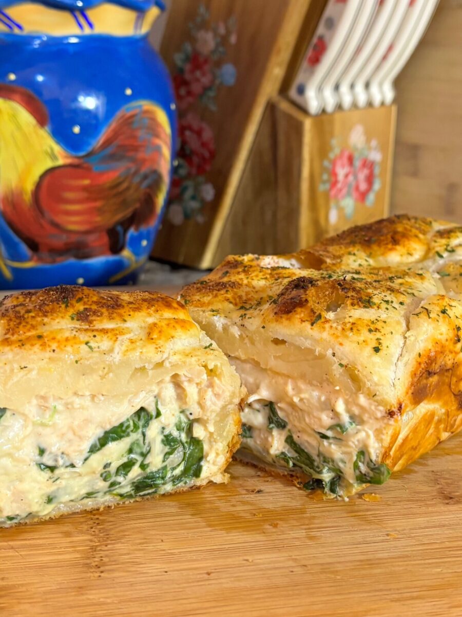 A close-up of a sliced puff pastry filled with creamy spinach and chicken, set on a wooden cutting board. In the background, there’s a colorful ceramic container with a rooster design and a holder with utensils.