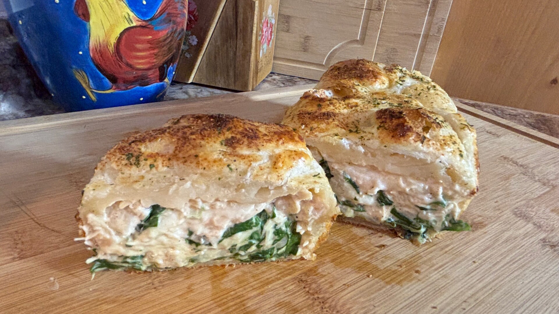 A homemade bread loaf sliced in half, revealing a creamy filling with spinach and herbs inside, sitting on a wooden cutting board with a colorful mug featuring a rooster in the background.