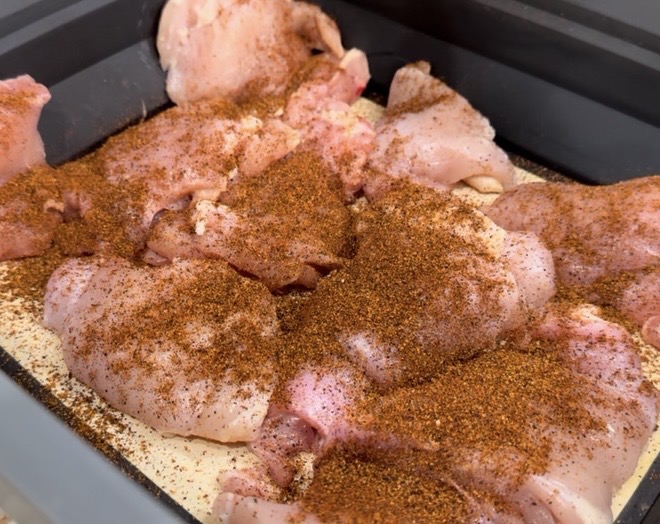 Raw chicken pieces in a black tray, generously coated with a reddish-brown spice mix, ready to be cooked.