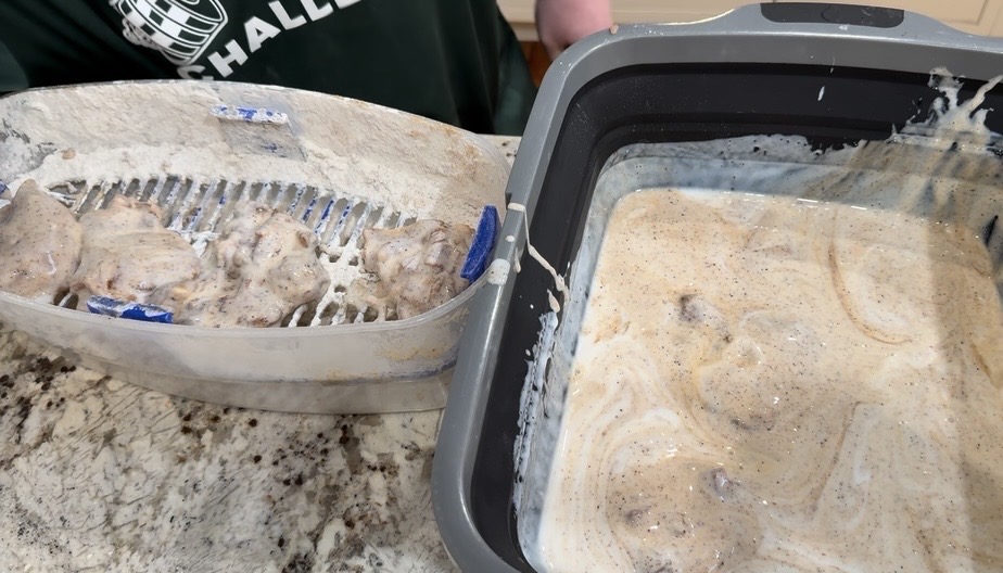 A person dips pieces of chicken in a creamy, seasoned batter. The battered chicken is placed on a rack over a tray next to a container filled with more batter, all on a granite countertop.