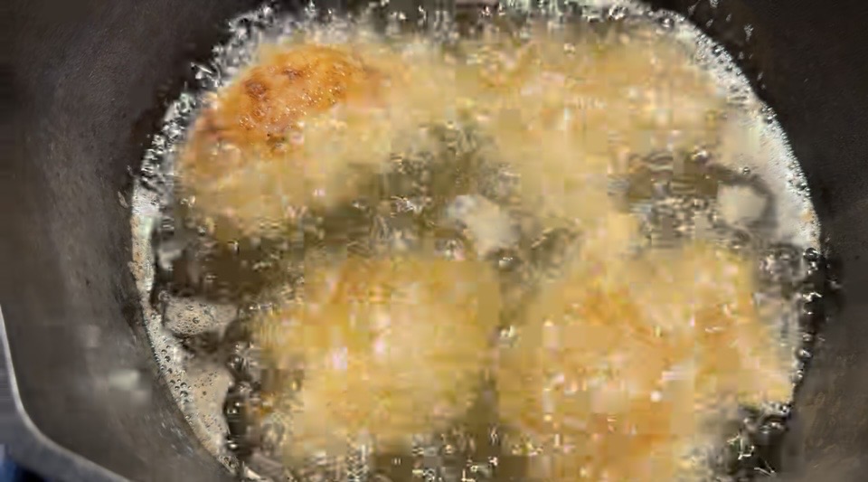 Four golden-brown round fritters are being deep-fried in bubbling hot oil inside a black pan.