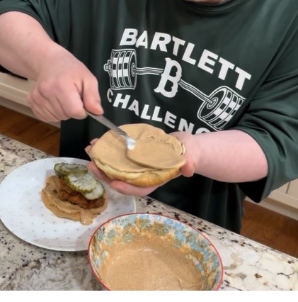 A person wearing a Bartlett Challenge shirt spreads peanut butter on a hamburger bun, with a plate holding a sandwich topped with pickles nearby and a bowl of peanut butter on the counter.